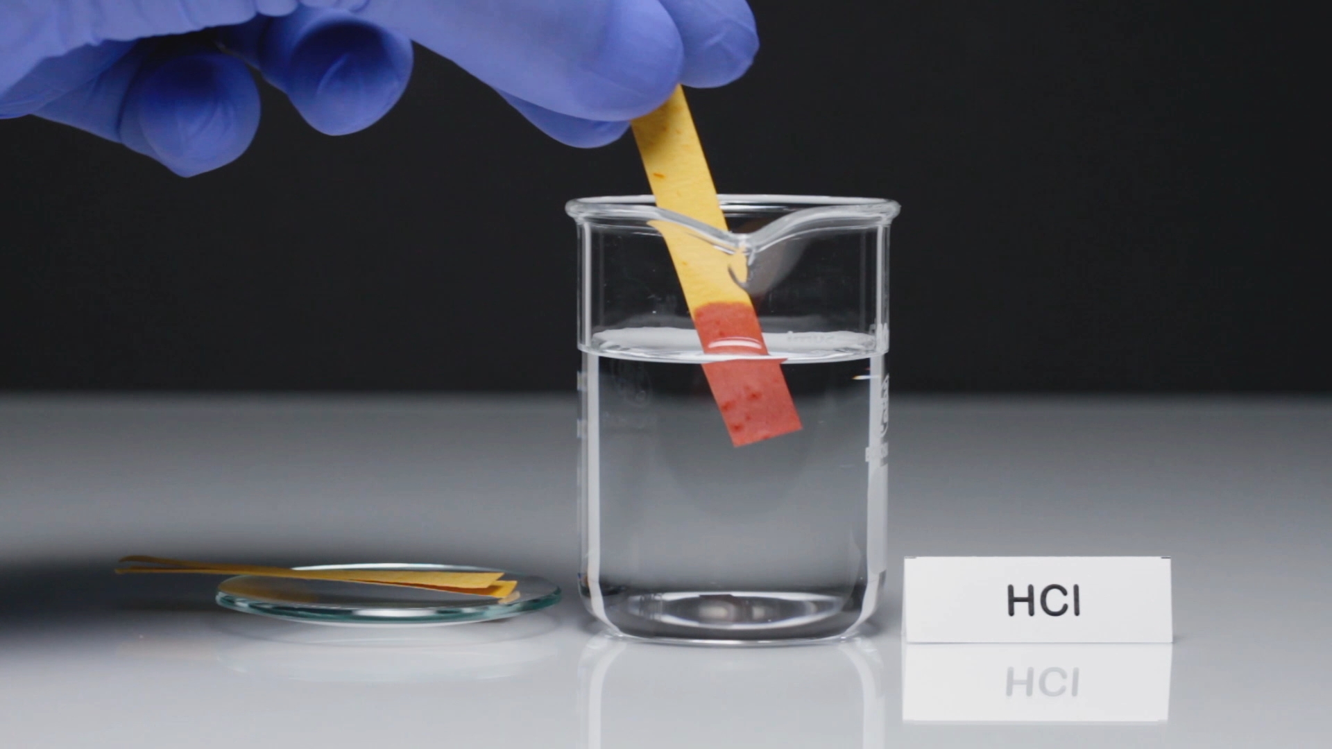 Ein Handschuhträger taucht einen pH-Teststreifen in ein Becherglas mit HCl-Lösung.