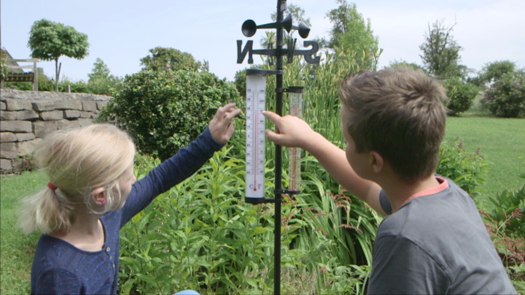 Ein Junge und ein Mädchen zeigen auf ein Thermometer im Garten, umgeben von Pflanzen.