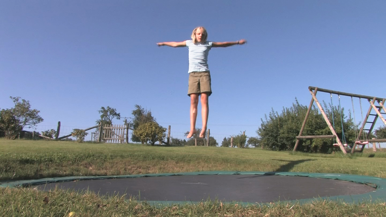 Ein Mädchen springt auf einem Trampolin im Freien, umgeben von Gras und Bäumen.