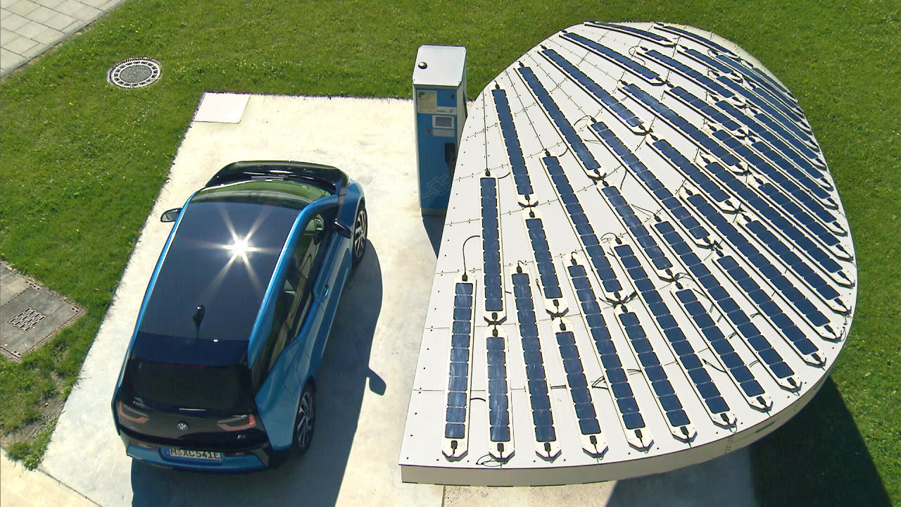 Ein blauer Elektroauto parkt unter einer Solaranlage mit geschwungener Form, die mit Solarpanelen bedeckt ist.