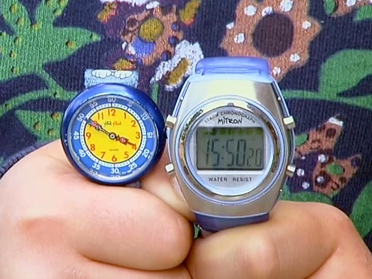 Eine Hand hält zwei Uhren: eine analoge Uhr mit blauen Ziffern und eine digitale Uhr mit grauem Gehäuse und blauer Armbanduhr.