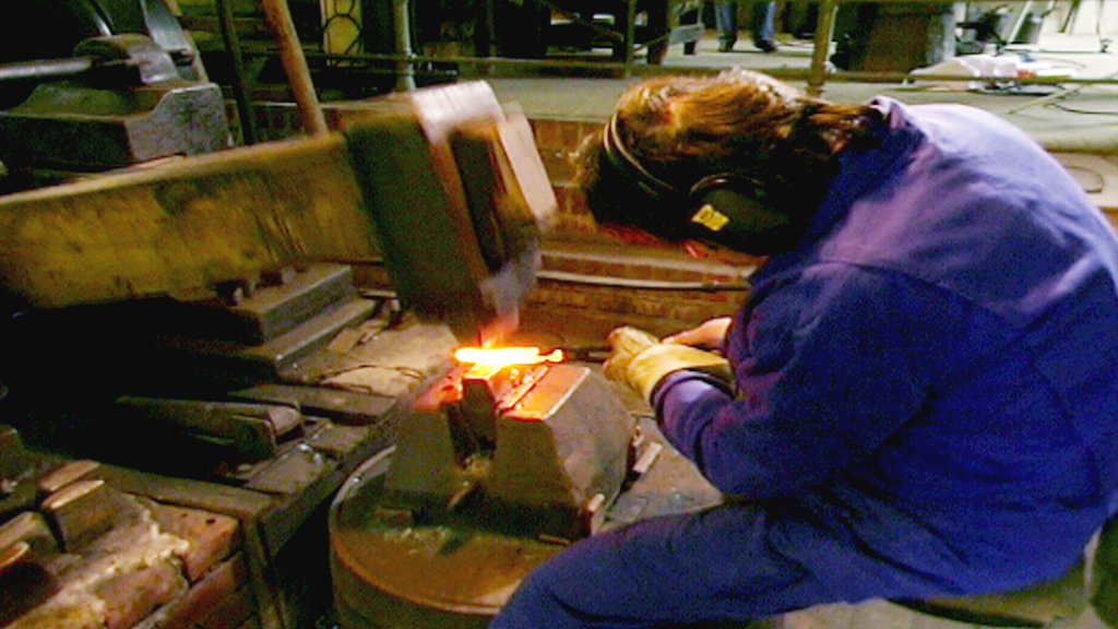 Ein Schmied arbeitet an einem Amboss, während ein Hammer auf ein glühendes Metallstück schlägt.