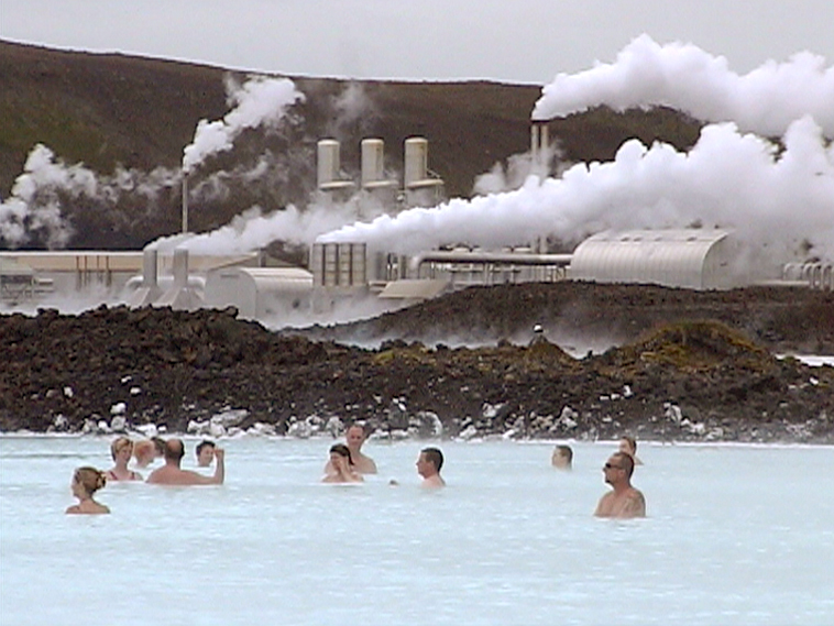 Menschen entspannen im blauen Wasser einer geothermalen Lagune, im Hintergrund dampfende Geothermiekraftwerke.