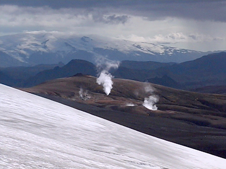 Dampfsäulen steigen aus der Erde in einer vulkanischen Landschaft mit schneebedeckten Bergen im Hintergrund.