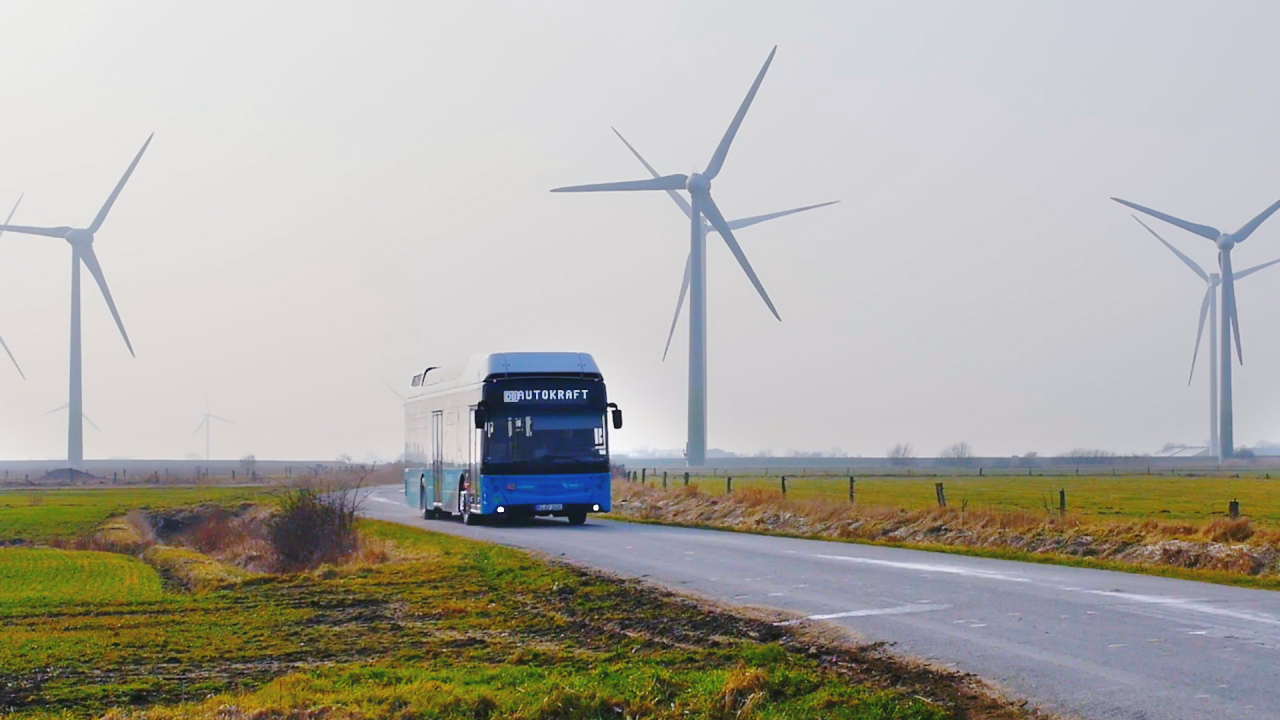 Ein blauer Reisebus fährt auf einer Straße zwischen Windkraftanlagen und Feldern.