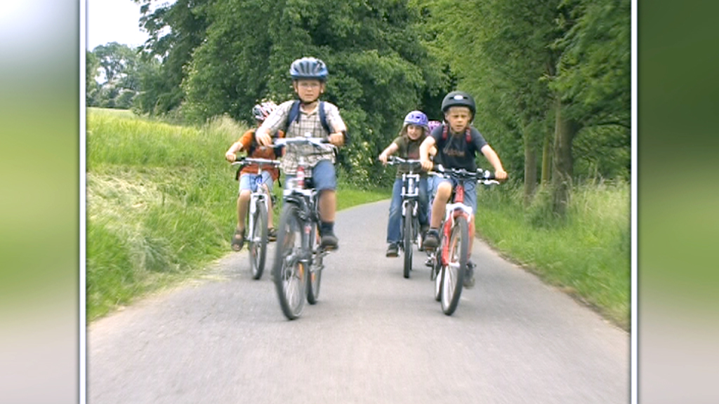 Fünf Kinder fahren auf Fahrrädern einen schmalen Weg entlang, umgeben von Bäumen und Wiese.