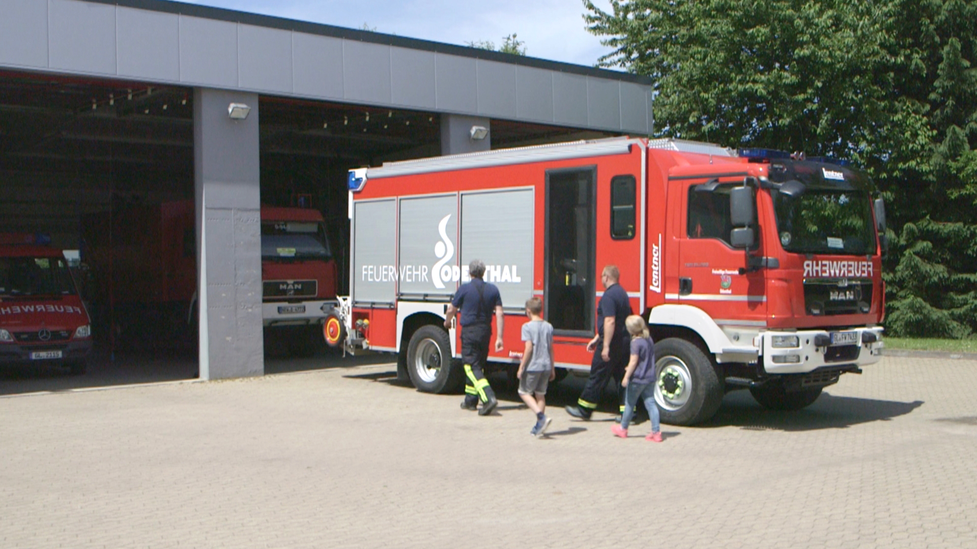 Ein rotes Feuerwehrfahrzeug steht vor einer Feuerwache, während Kinder in Richtung des Fahrzeugs gehen.
