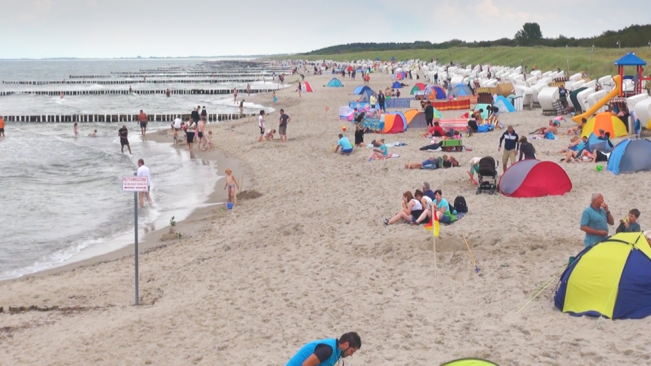 Menschen am Strand mit bunten Zelten, Wellen und Holzstegen im Hintergrund.