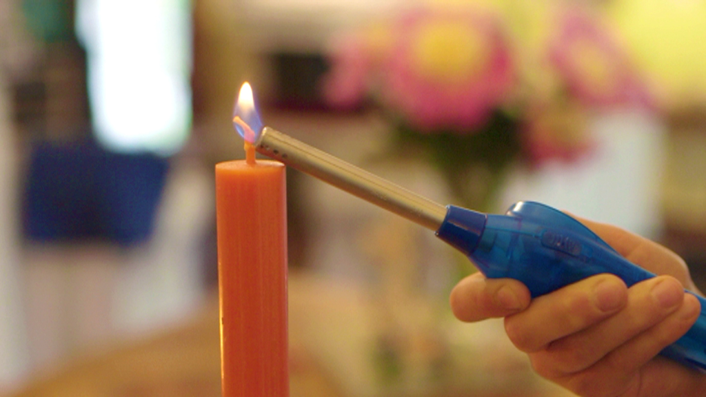 Eine Hand hält einen langen Feuerzeug, um eine orangefarbene Kerze anzuzünden.