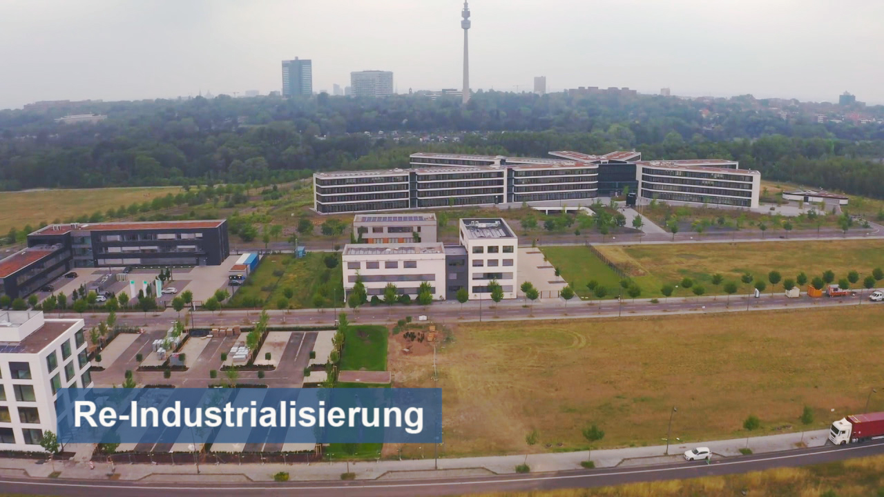 Luftaufnahme eines modernen Industriegebiets mit mehreren Gebäuden und einem Fernsehturm im Hintergrund.