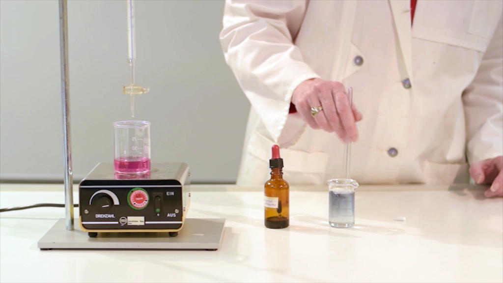 Ein Wissenschaftler in einem weißen Laborkittel führt ein Experiment durch, indem er eine Pipette in ein Glas mit blauer Flüssigkeit taucht.