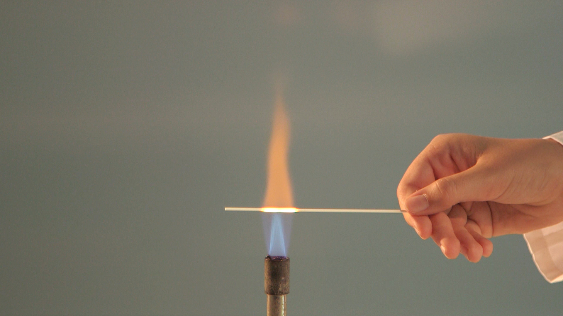 Eine Hand hält einen Metallstab über einer Flamme, die orange und blau leuchtet.