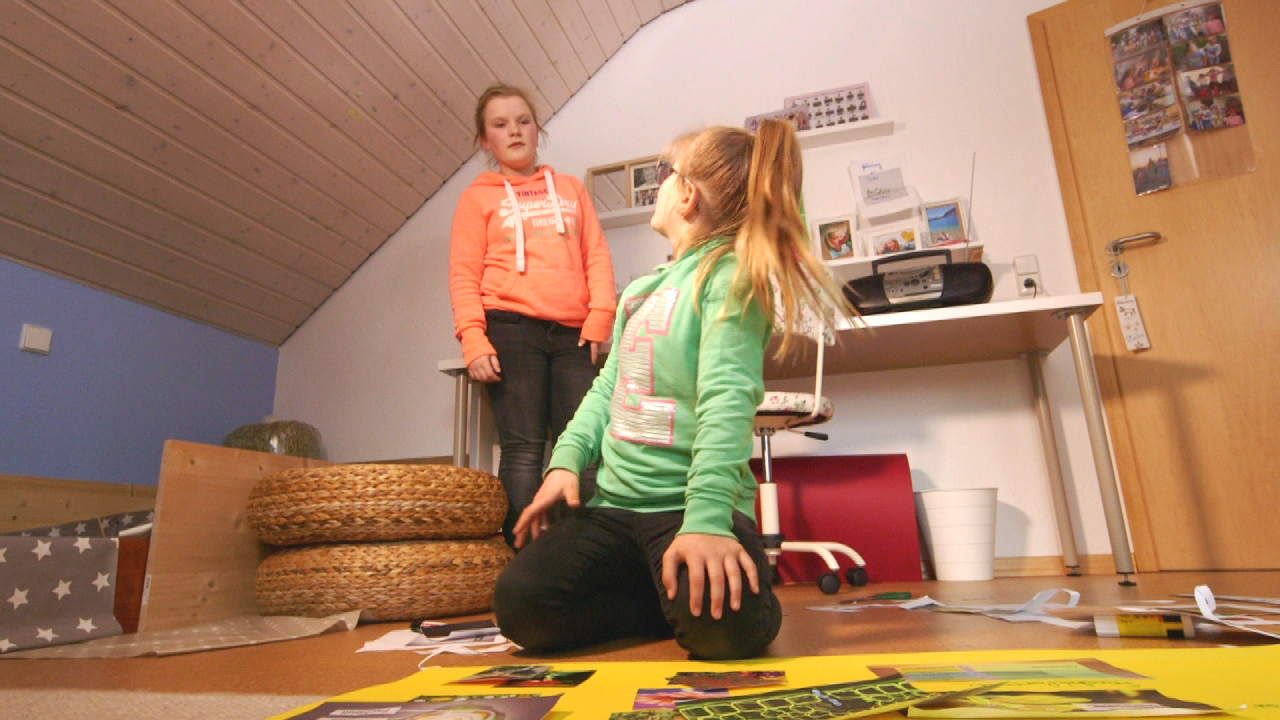 Zwei Mädchen in einem Zimmer, eines steht und schaut das andere an, das auf dem Boden sitzt und mit Bastelmaterial arbeitet.