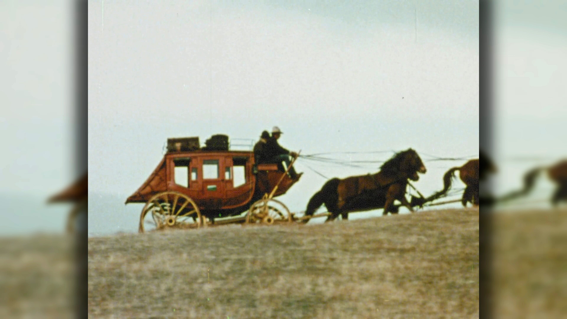 Eine historische Postkutsche wird von zwei Pferden über eine hügelige Landschaft gezogen.