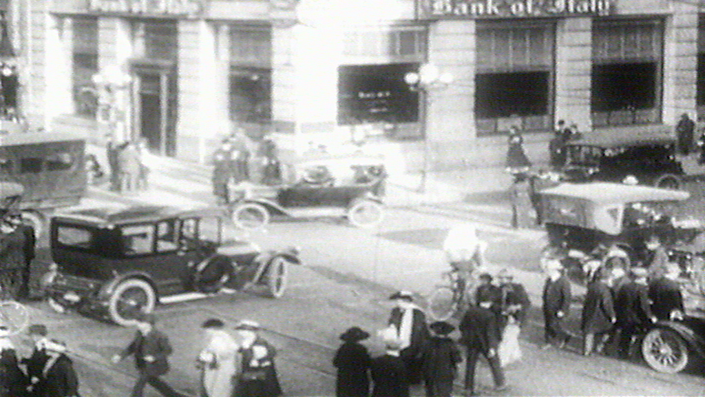 Historische Straßenszene mit Autos, Fußgängern und einem Bankgebäude im Hintergrund.