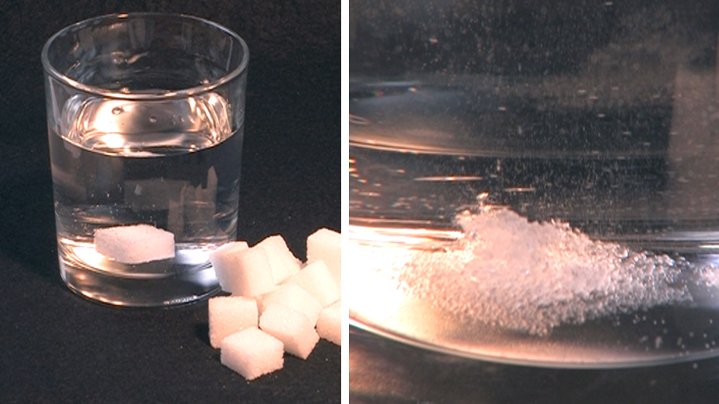 Ein Glas Wasser mit Zuckerstücken daneben. Auf der rechten Seite ist Zucker, der sich im Wasser auflöst.