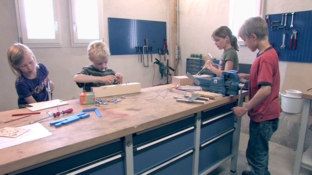 Vier Kinder arbeiten in einer Werkstatt, zwei stehen am Tisch und basteln, während die anderen an einem Werkbank stehen.