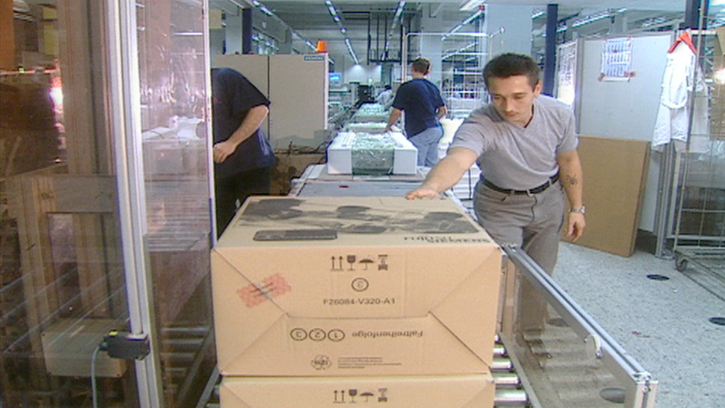 Ein Mann in grauem Shirt, der eine Kiste auf einem Förderband in einer Fabrik anfasst. Im Hintergrund sind weitere Arbeiter zu sehen.
