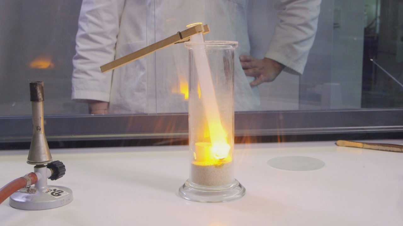 Ein Experiment mit einer Flamme in einem Glasbehälter, der auf einem Tisch steht. Im Hintergrund ist eine Person in einem Laborkittel zu sehen.