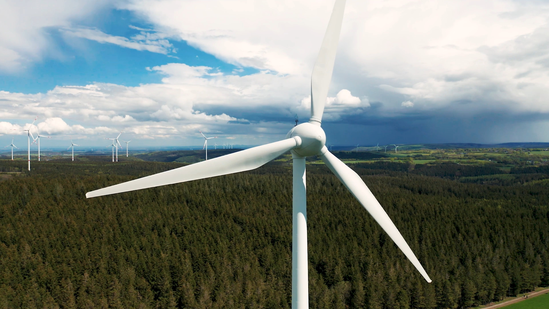 Nahaufnahme eines weißen Windrads vor bewaldeter Landschaft und bewölktem Himmel.