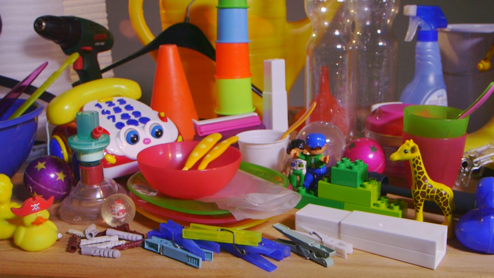 Bunte Kinderspielzeuge und Haushaltsgegenstände wie Bauklötze, Giraffenfigur, Telefon, Schüsseln, Wäscheklammern und Sprühflasche auf einem Tisch.