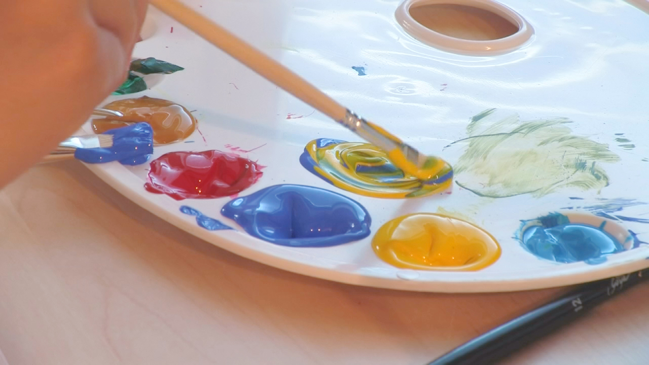 Eine Hand hält einen Pinsel und mischt gelbe und grüne Farbe auf einer Paletten.