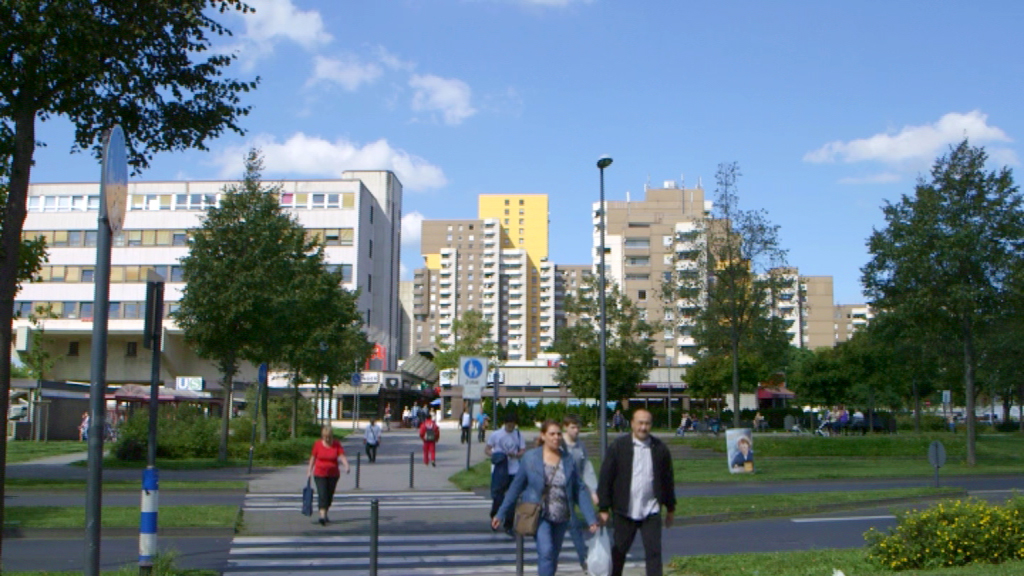 Stadtansicht mit mehreren Wohngebäuden und Fußgängern auf einem Gehweg.