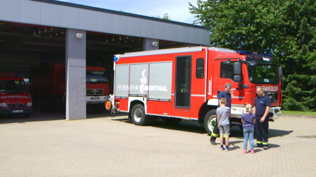 Feuerwehrfahrzeug vor der Feuerwache mit zwei Feuerwehrleuten und zwei Kindern.
