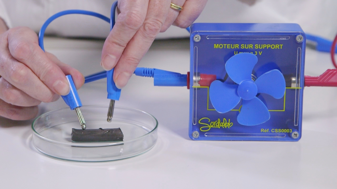 Ein blauer Motor mit einem Ventilator, der an eine Platte angeschlossen ist, während eine Hand die elektrischen Anschlüsse hält.
