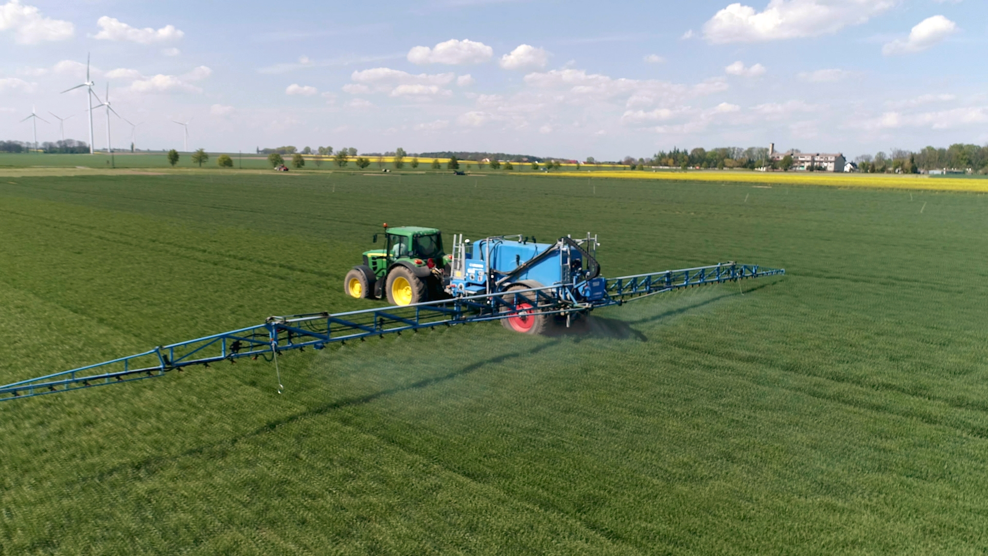 Traktor mit breitem Anbaugerät fährt über ein großes Feld mit grünem Getreide.