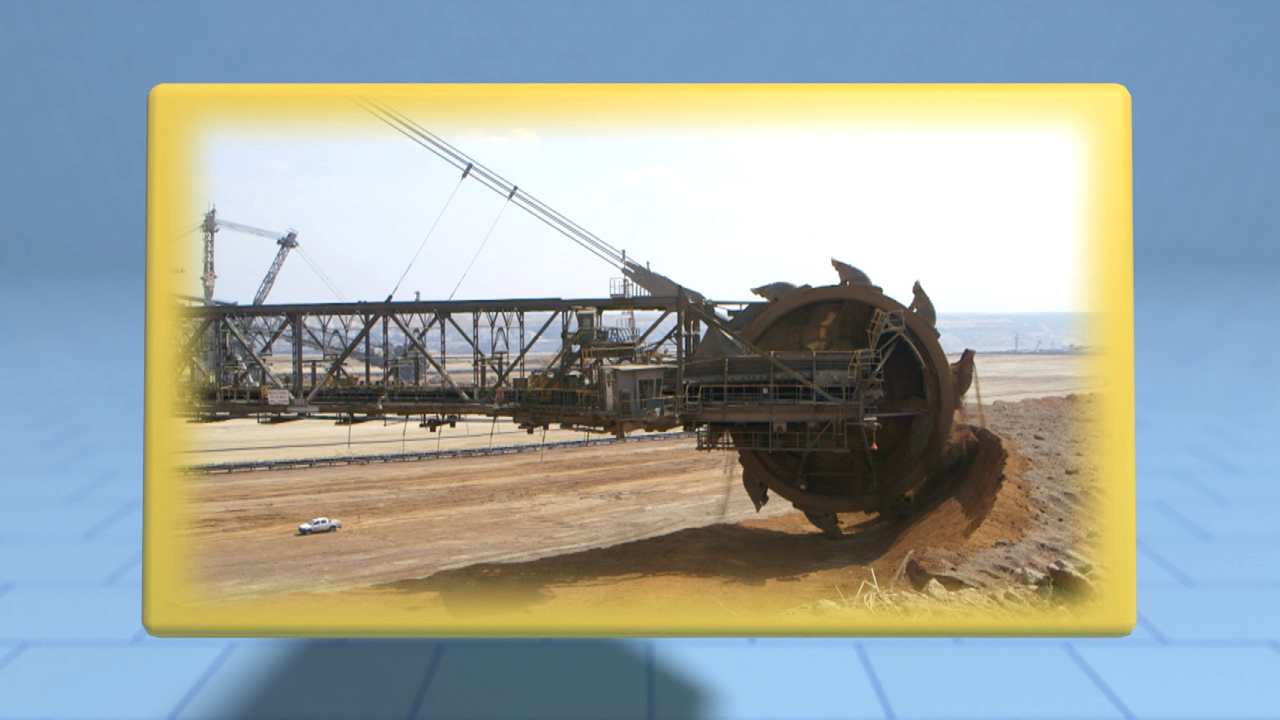 Ein großer Bagger im Tagebau, der Erde abträgt. Im Hintergrund sind Baukräne zu sehen.