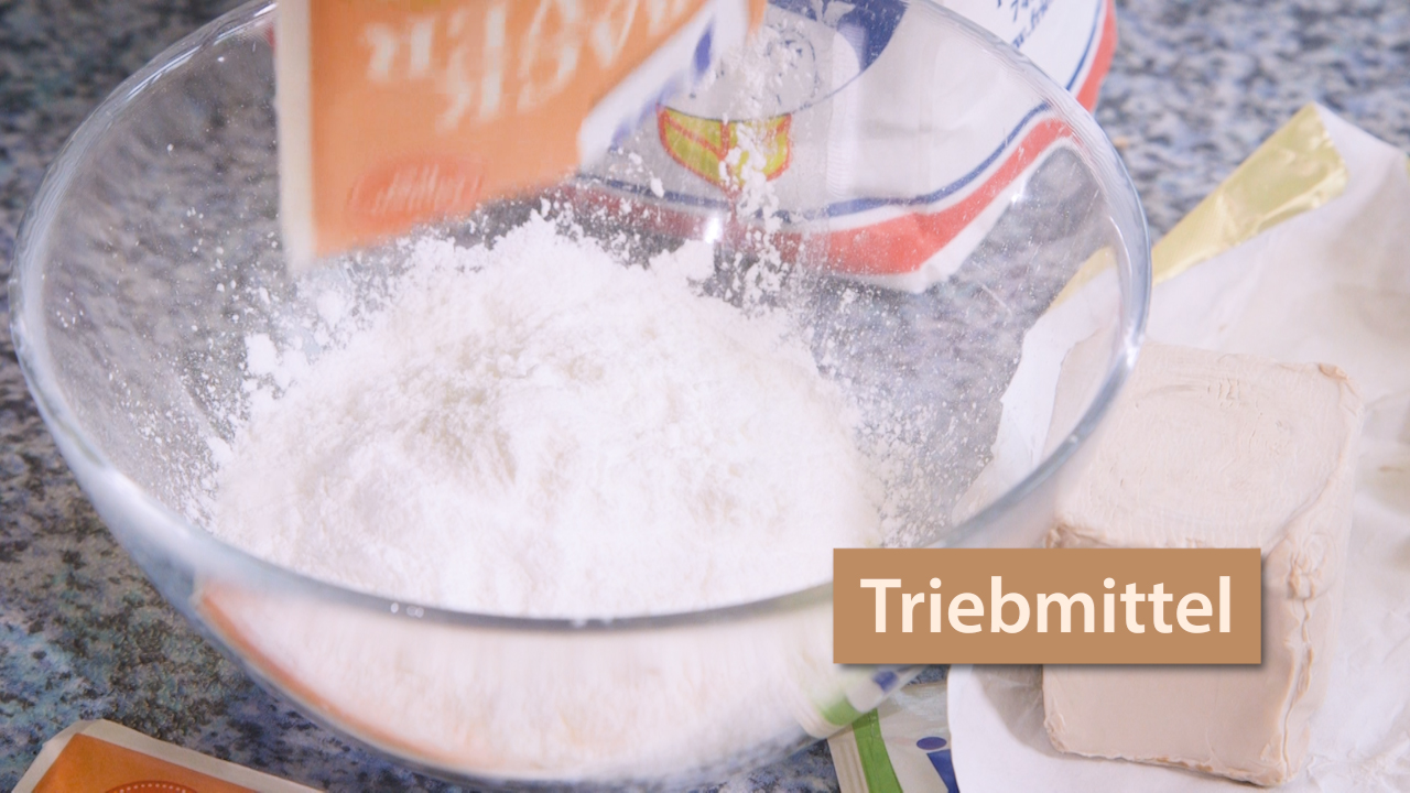 Eine Schüssel mit Mehl, in die Triebmittel eingestreut wird. Im Hintergrund ist ein Stück Hefe zu sehen.