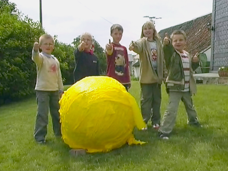 Fünf Kinder stehen im Freien und zeigen mit Daumen nach oben vor einem großen gelben Ball aus Plastikfolie.