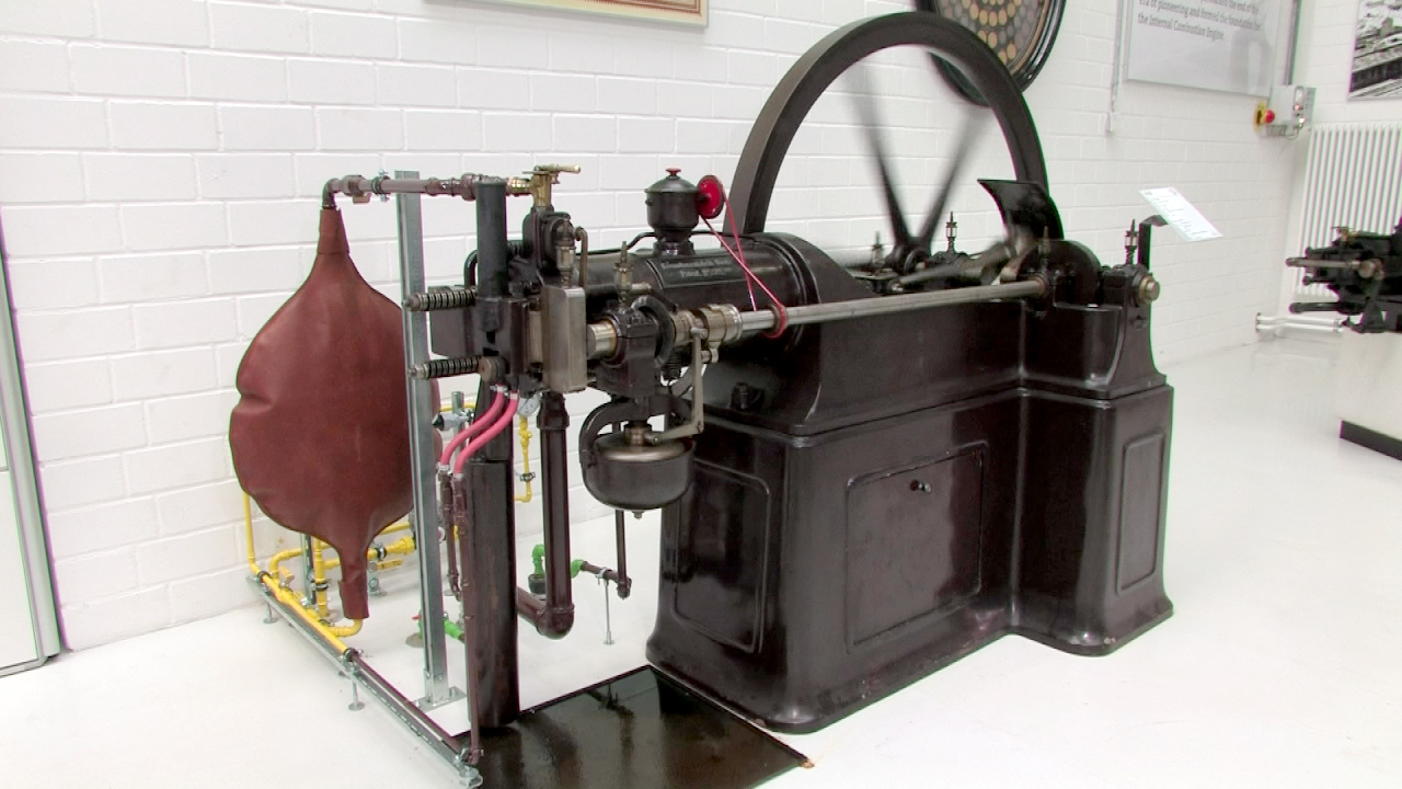 Dampfmaschine mit großen Schwungrad und verschiedenen Rohren, in einem modernen Ausstellungsraum.