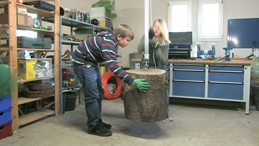 Zwei Kinder heben einen Baumstamm in einer Werkstatt, umgeben von Werkzeugen und Materialien.