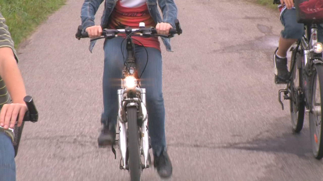 Drei Personen fahren auf Fahrrädern auf einer Straße. Eine Person in der Mitte fährt ein weißes Fahrrad mit einem Licht.