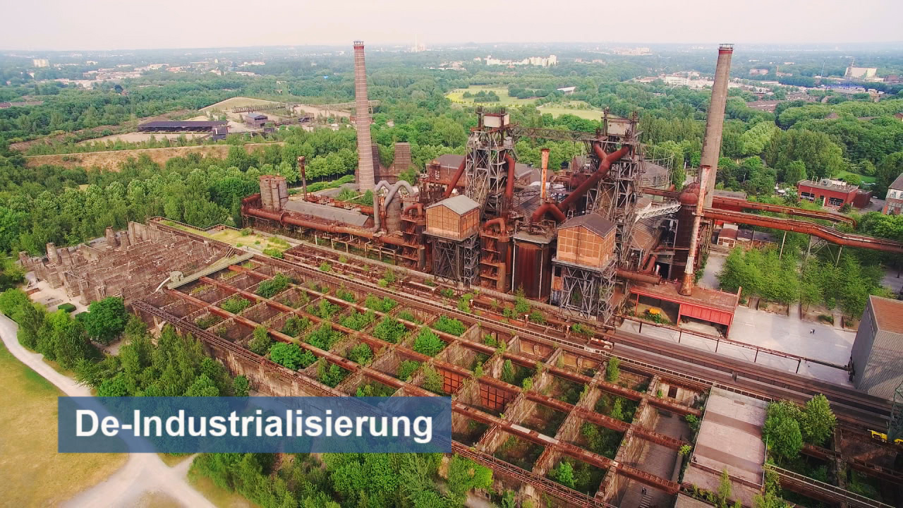 Luftaufnahme einer stillgelegten Industrieanlage mit rauchenden Schornsteinen und grünen Flächen im Vordergrund.