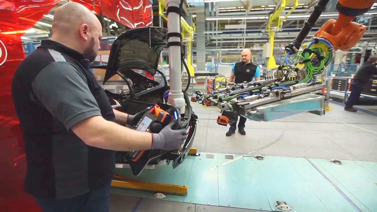 Ein Arbeiter hält ein Autoteil in der Hand, während ein Roboterarm ein anderes Teil anhebt. Im Hintergrund ist eine Produktionslinie zu sehen.