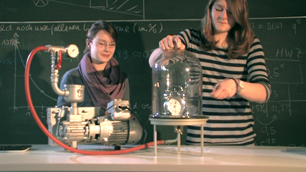 Zwei Frauen führen ein Experiment mit einer Vakuumpumpe und einer Glaskuppel durch, während sie an einer Tafel mit mathematischen Formeln stehen.