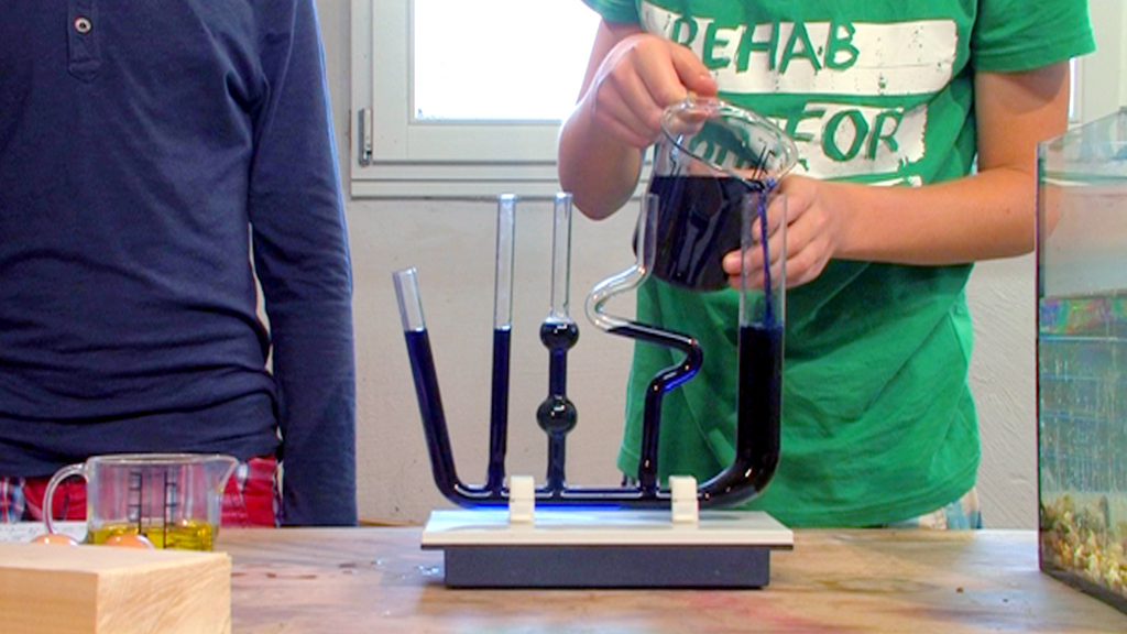Zwei Kinder führen ein Experiment mit einem Apparatur durch, während eine Flüssigkeit in ein Gefäß gegossen wird.