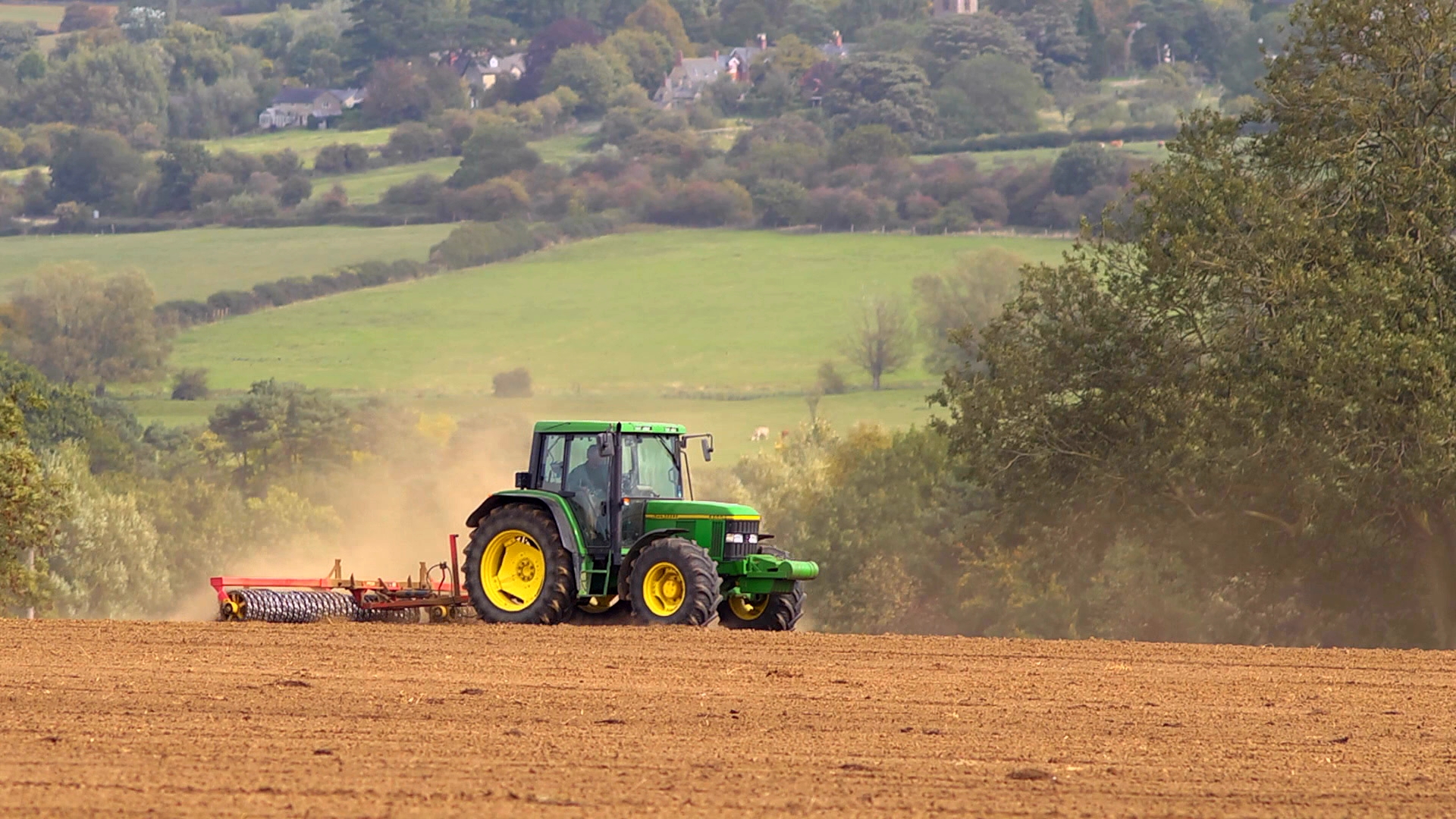 Traktor bearbeitet ein großes, frisch gepflügtes Feld vor einer grünen Landschaft mit Bäumen und Hügeln.