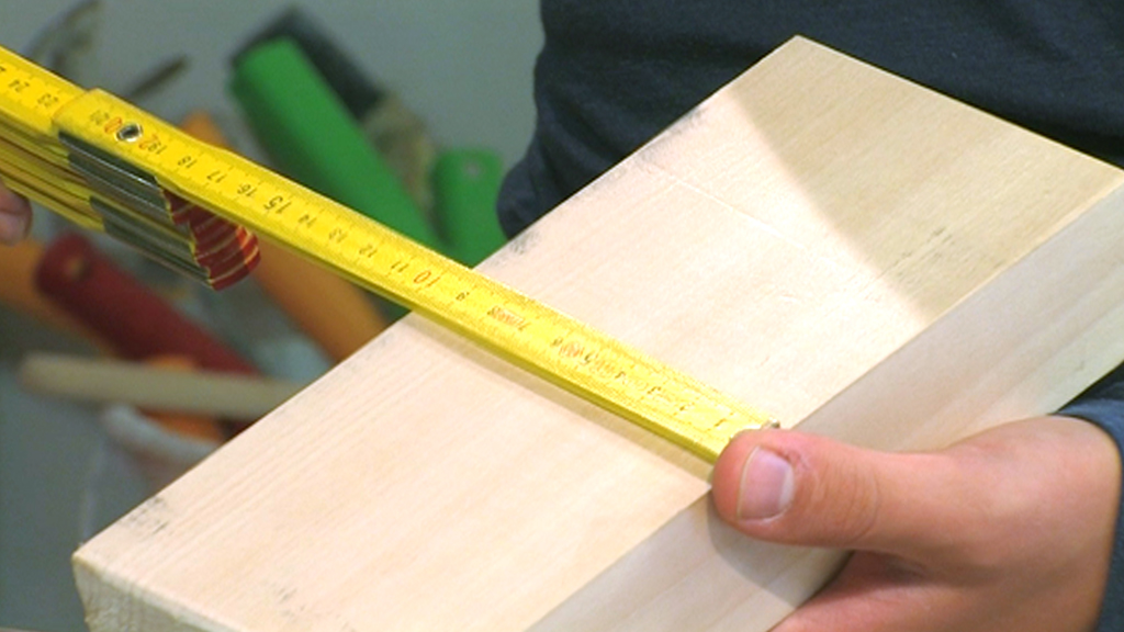 Eine Person hält ein Stück Holz und misst es mit einem gelben Zollstock.