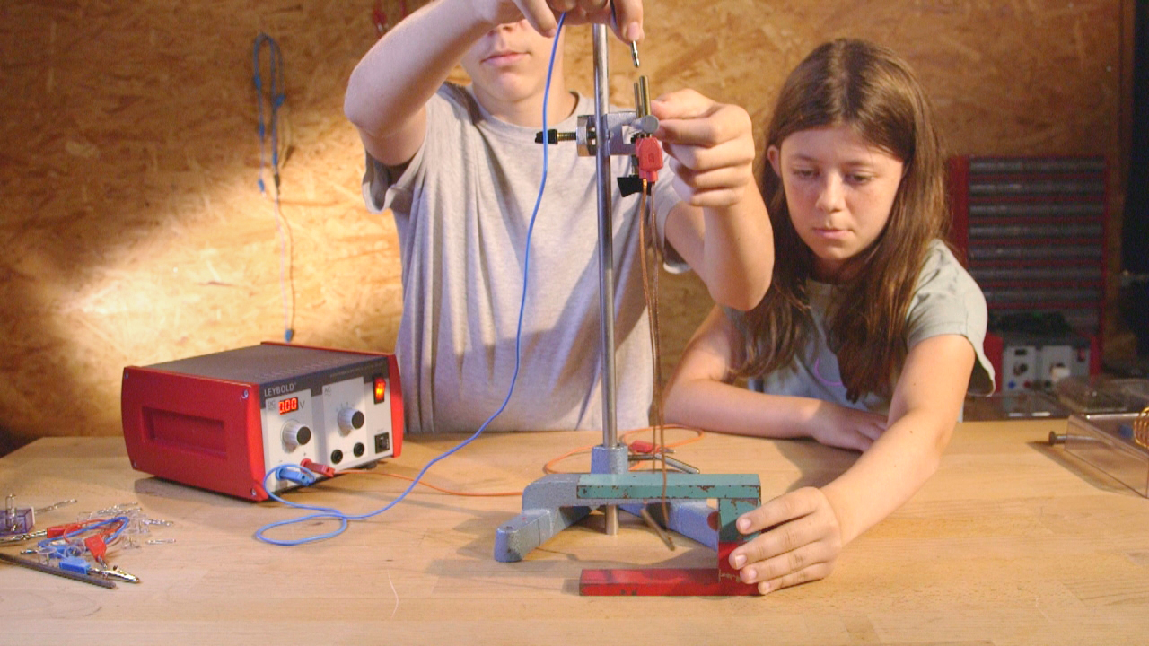 Zwei Kinder führen ein Experiment durch, indem sie ein Gerät justieren. Das Mädchen beobachtet aufmerksam, während der Junge die Einstellungen vornimmt.
