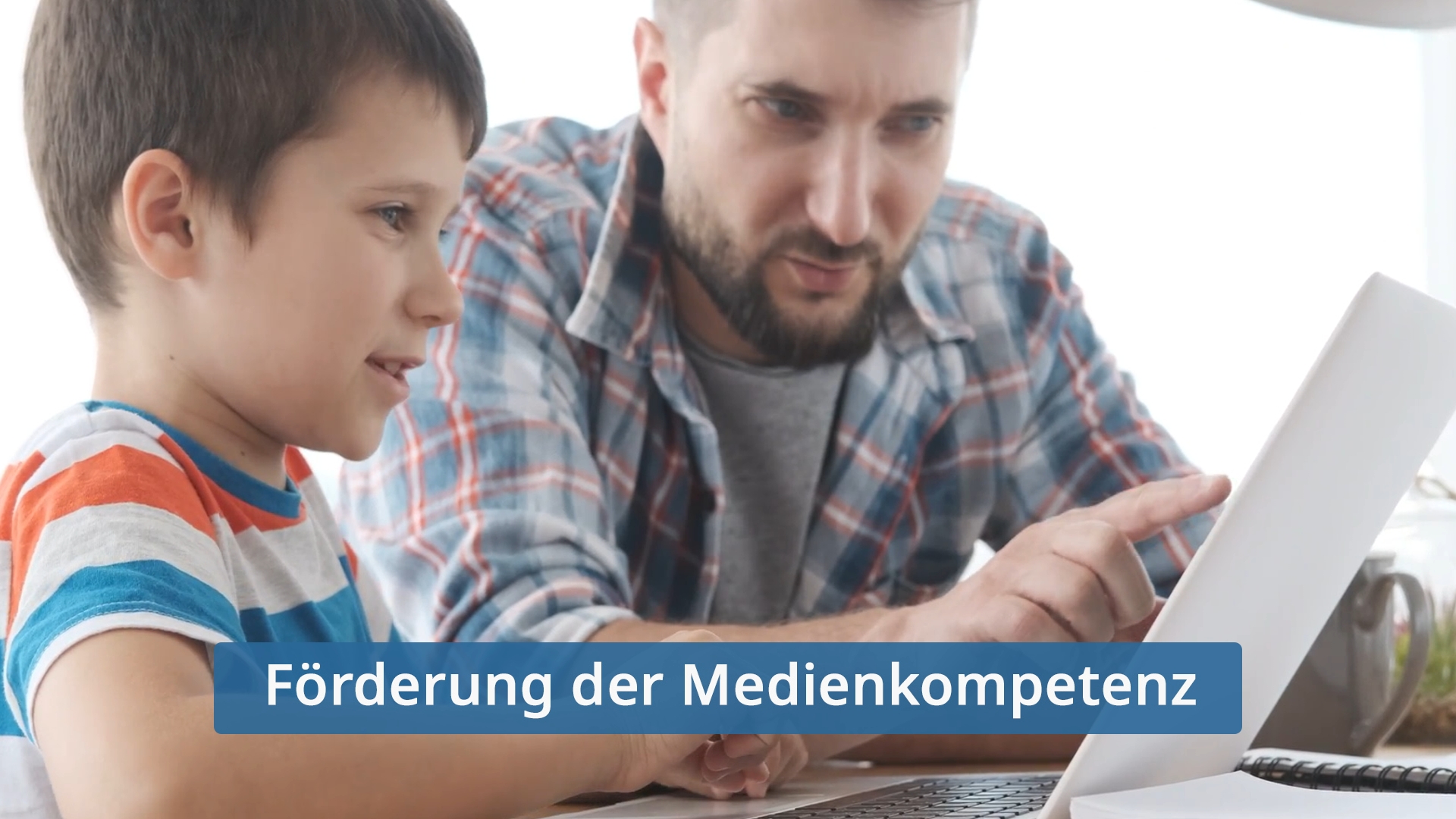 Ein Junge und ein Mann sitzen an einem Tisch mit einem Laptop. Der Junge trägt ein gestreiftes T-Shirt, der Mann ein kariertes Hemd. Text: 'Förderung der Medienkompetenz'.