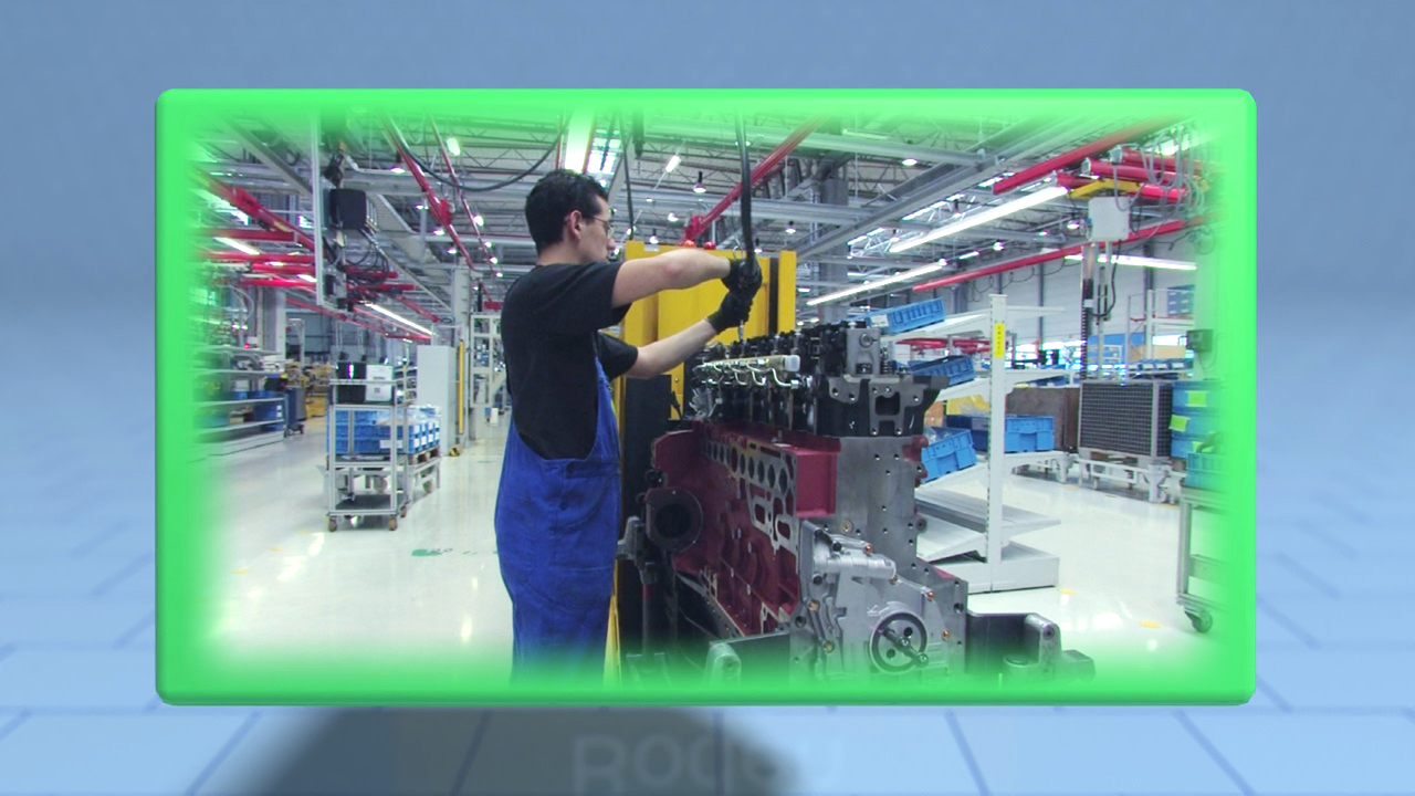 Ein Arbeiter in blauer Latzhose montiert einen Motorblock in einer modernen Fabrik.