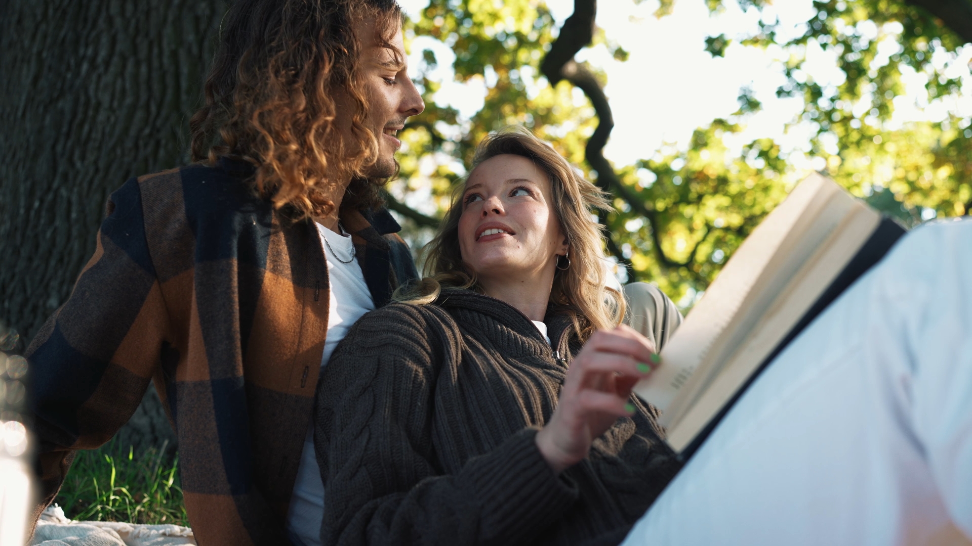 Ein Paar sitzt im Freien unter einem Baum. Der Mann mit lockigem Haar schaut die Frau an, die ein Buch hält und lächelt.