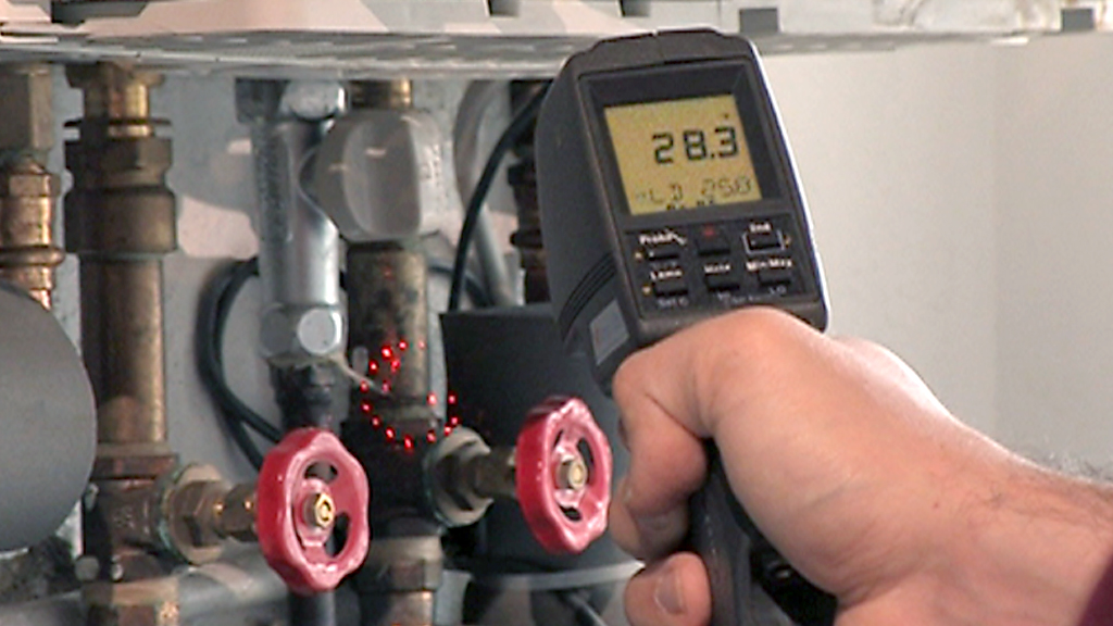 Eine Person misst die Temperatur mit einem digitalen Thermometer an einer Heizungsanlage mit roten Ventilen.