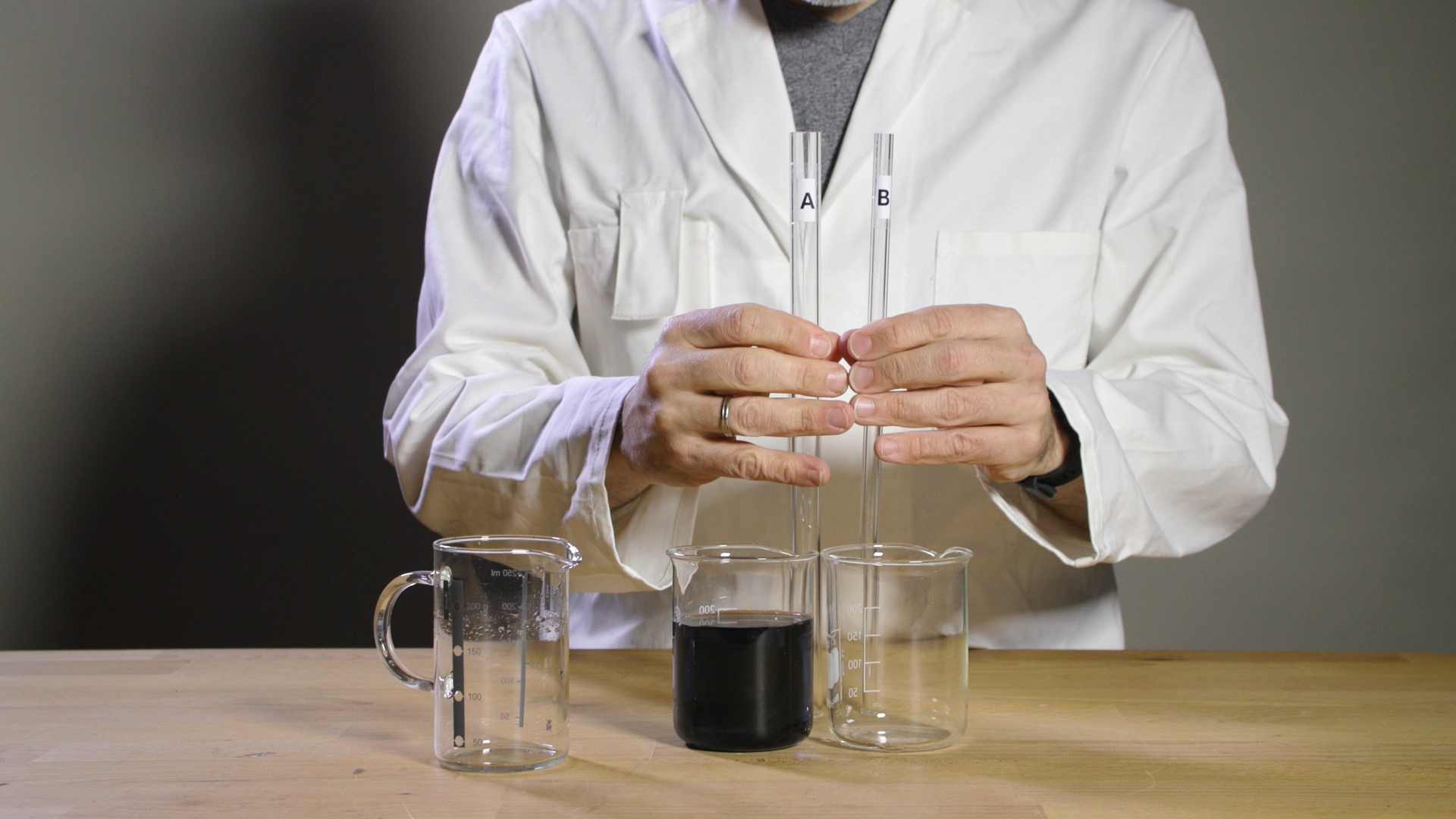 Person in weißem Laborkittel hält zwei Reagenzgläser mit den Aufdrucken 'A' und 'B' über drei Bechergläser auf einem Holztisch, davon eines mit schwarzer Flüssigkeit gefüllt