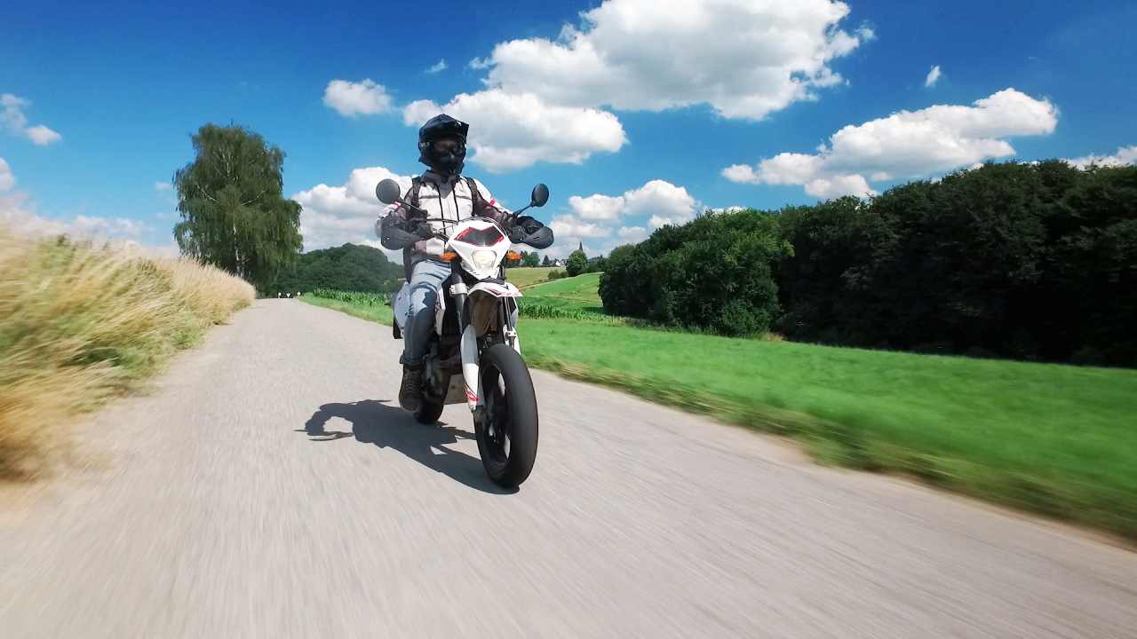 Motorradfahrer auf einer Landstraße, umgeben von grünen Feldern und Bäumen unter blauem Himmel.