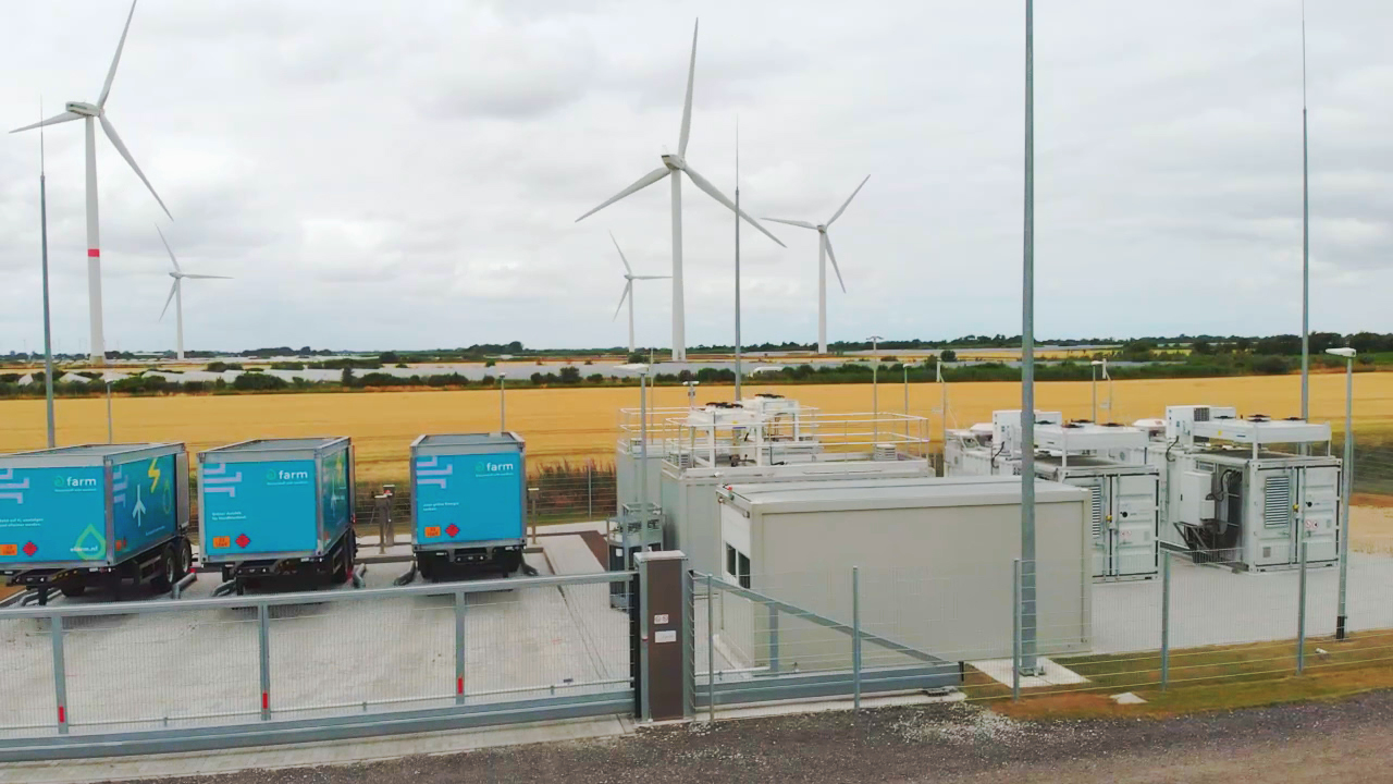 Energieanlage mit mehreren blauen Containern und Windkraftanlagen im Hintergrund.